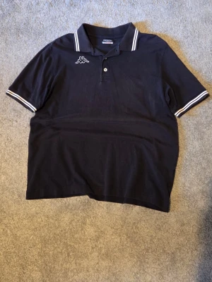 Svart pikétröja från Kappa - Svart pikétröja från Kappa med vit logga på bröstet och vita ränder på krage och ärmslut. Klassisk polokrage med knappar framtill. Perfekt för en sportig och avslappnad stil.