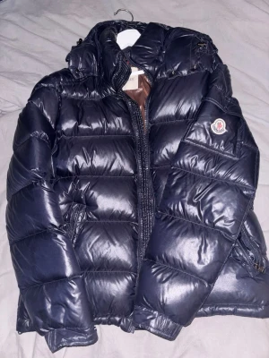 Svart dunjacka från Moncler - Snygg blå dunjacka från Moncler med glansig finish och brunt innerfoder. Jackan har huva, dragkedja framtill, två sidofickor med dragkedja och Moncler-logga på ärmen. Perfekt för kalla vinterdagar och riktigt varm tack vare dunfyllningen.