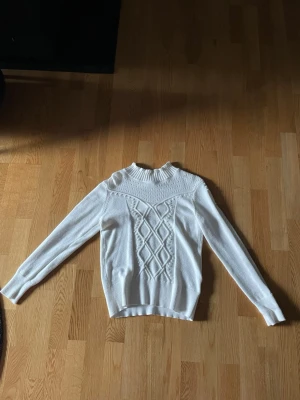 Vit stickad tröja från Stories - Supersnygg vit stickad tröja från Stories med ribbad hög hals och detaljerat mönster framtill. Tröjan har långärmade ärmar med strukturdetaljer längs sidorna och ribbade muddar. Perfekt för dig som gillar en clean men ändå unik look.