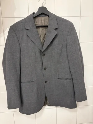 Grå kavaj från Tiger of Sweden - Stilren grå kavaj från Tiger of Sweden med klassisk passform och tre knappar framtill. Kavajen har två fickor med lock och en bröstficka. Fodrad insida och snyggt snitt, perfekt för en dressad look.