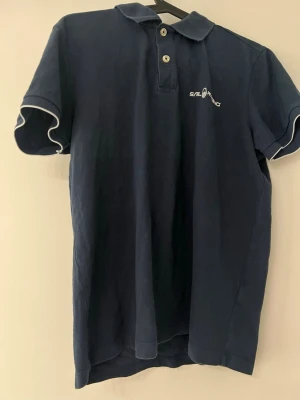 Marinblå pikétröja från Sail Racing - Klassisk marinblå pikétröja från Sail Racing med vit logga på bröstet och vita detaljer vid ärmsluten. Tröjan har krage, knappar framtill och korta ärmar. Perfekt till en avslappnad och sportig stil.