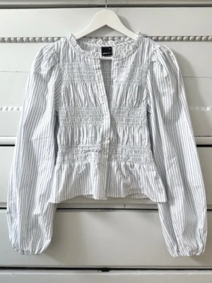 Randig blus  - Snygg blus från Gina Tricot i vitt med tunna grå ränder. Blusen har smockdetaljer över bröst och rygg, v-ringning och långa puffärmar med resår vid ärmslut. Tillverkad i ett lätt och luftigt tyg, perfekt för sommaren. priset går att diskutera💕