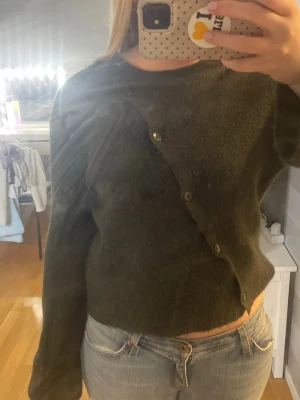 Mörkgrön asymmetrisk kofta - Säljer en mörkgrön kofta med asymmetrisk knäppning framtill. Koftan har långa ärmar och är gjord i ett mjukt, stickat material. Perfekt för dig som gillar unika detaljer och en avslappnad stil.