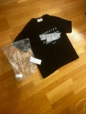 Svart Maison Margiela t-shirt - Säljer en svart t-shirt från Maison Margiela med det ikoniska numermotivet tryckt på framsidan . Materialet är mjuk bomull och passformen är normal. Perfekt för dig som gillar stilrena och trendiga plagg. Pris är diskuterbart!!