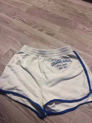 Vita shorts med blå kant från H&M - Säljer ett par vita shorts från H&M med blå kant längs nederkanten. De har elastisk midja och är tillverkade i mjuk bomull, perfekta för en avslappnad stil. Passar dig som gillar sportiga och bekväma plagg.