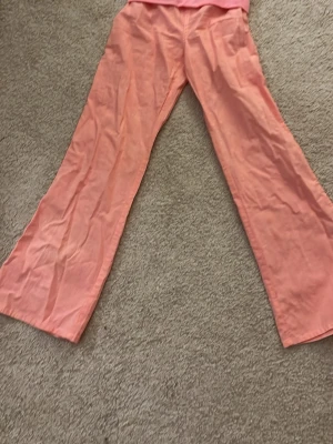 Virala lågmidjade korall linnebyxor - Säljer ett par vida linnebyxor i en snygg korall färg. Byxorna har låg midja och utsvängda ben, perfekta för en chill och avslappnad stil. Tillverkade i ett tunt och luftigt linnetyg som känns skönt mot huden.
