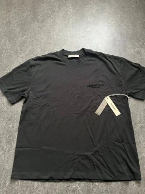  Essentials t-shirt - Snygg svart t-shirt från Essentials Fear of God med diskret logga på bröstet och större tryck på ryggen. Klassisk passform och rund hals. Tillverkad i mjuk bomull som känns skön mot huden. Perfekt för dig som gillar streetwear och stilrena plagg.
