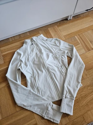 Lululemon Define Jacket Nulu - Snygg vit sportjacka från Lululemon med hög krage och dragkedja framtill. Jackan har två fickor med dragkedja, långa ärmar och tumhål vid muddarna. Tillverkad i ett stretchigt och mjukt material som passar perfekt till träning eller promenader. Nypris 1380kr