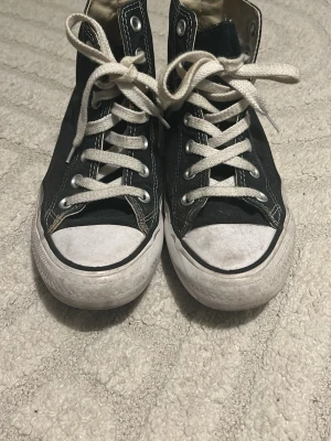 Svarta Converse All Star Highs - Klassiska svarta Converse Chuck Taylor All Star med högt skaft och vita gummisulor. Skorna har vita snören, metallöljetter och den ikoniska loggan på sidan. Ovandelen är i slitstark canvas och sulan är i gummi. Perfekta för en avslappnad streetstil.