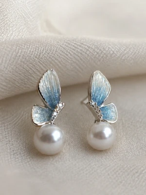 Blå fjärilsörhängen med pärlor - Dessa örhängen inspirerade av fairy wings i morgonljus – skimrande, lätta och eleganta. De fina vingarna fångar ljuset med ett mjukt glitter, medan pärlan ger en tidlös och balanserad finish.                                   Material: Stift i 925 sterlingsilver (stämplat), detaljer i rostfritt stål.                    Nickeltestat & allergivänlig.                 Storlek : längd 3 cm Bredd 1 cm