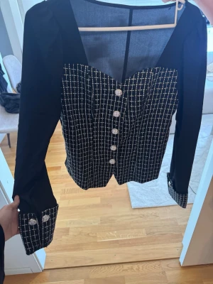 Svart blus med rutigt mönster och pärlknappar - Stilren svart blus med lång ärm och rutigt mönster i vitt framtill och på manschetterna. Blusen har dekorativa pärlknappar och en markerad midja med spetsig nederkant. Perfekt för dig som gillar en chic och trendig look.