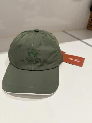Olivgrön keps från Loro Piana  - Snygg olivgrön keps från LP med broderad logga framtill. Klassisk böjd skärm och justerbar passform bak. Tillverkad i ett lätt och slitstarkt material som passar perfekt till streetwear eller casual outfits.