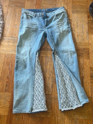 Unika ljusblå jeans med spets - Säljer ett par ljusblå jeans från Z1975 med breda ben och insydd vit spets i slitsarna framtill köpta för 1000kr handsydda. Spetsjeans lowrise Lågmidjade utsvängda bootcut baggy lowwaist.Jeansen har klassisk femficksmodell och knappgylf. Perfekta för dig som vill sticka ut med en kreativ och personlig stil. Spetsjeans lowrise Lågmidjade utsvängda bootcut baggy lowwaist