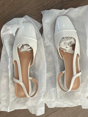 Vita ballerinaskor med rem och låg klack - Det här är vita sandaler/klackskor, helt nya, oanvända alls, ligger fortfarande kvar i väskan och papperet ligger fortfarande i dem. De har en liten klack. De här var lite små i storleken, så tänk på det. Jag brukar ha storlek 39, men de här är små i storleken, så den här kanske passar bra om du har storlek 37-38.