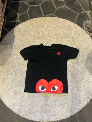 Svart Comme des Garçons Play t-shirt - Svart t-shirt från Comme des Garçons Play med det ikoniska röda hjärtat på bröstet och ett stort rött hjärta med ögon längst ner framtill. Klassisk passform och rund hals. Perfekt för dig som gillar streetwear och vill sticka ut.