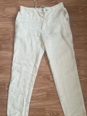 Vita linnebyxor med dragsko - Säljer ett par vita linnebyxor med dragsko i midjan och knappgylf. Byxorna har raka ben, sidfickor fram och två bakfickor med knapp. Perfekta för en avslappnad och stilren look. Materialet är svalt och luftigt linne.