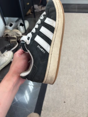Adidas Campus svarta sneakers - Klassiska Adidas Campus sneakers i svart mocka med vita läderdetaljer och de ikoniska tre ränderna på sidan. Skorna har vita skosnören och en beige sula som ger en retrovibe. Perfekta för dig som gillar streetwear och tidlös stil. Box och nya snören finns 