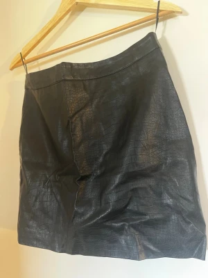 Svart kortkjol i skinnimitation - Säljer en svart kortkjol i skinnimitation med croco-mönstrad yta. Kjolen har en stilren och trendig look som passar perfekt till alla tillfällen. 