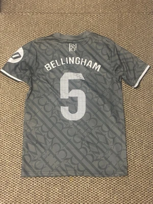 Real Madrid Bellingham matchtröja - Snygg grå matchtröja från Real Madrid med Bellingham och nummer 5 på ryggen. Tröjan har vita detaljer, klubbmärke på bröstet och Emirates Fly Better som sponsortryck. Tillverkad i lätt och ventilerande material, perfekt för fotboll eller träning.