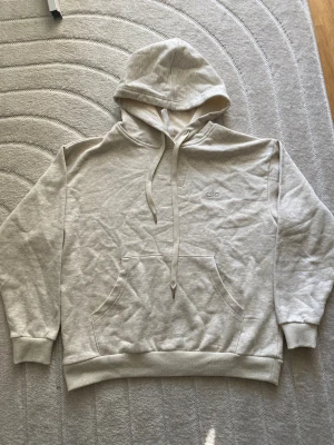 Beige hoodie från Alo med huva - Säljer en beige hoodie från Alo med broderad logga på bröstet. Hoodien har stor känguruficka framtill, dragsko i huvan och ribbade muddar vid ärmar och nederkant. Tillverkad i mjukt bomullsmaterial för en avslappnad look.