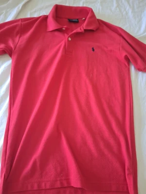 Rosa pikétröja från Ralph Lauren - Klassisk rosa pikétröja från Ralph Lauren med broderad logga på bröstet. Tröjan har krage, korta ärmar och knäppning med två knappar framtill. Perfekt för en stilren och sportig look. Varan är i mycket bra skick i storlek M slim fit. Hör av dig vid frågor