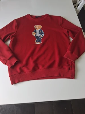 Röd sweatshirt Polo Ralph Lauren - Röd sweatshirt från Polo Ralph Lauren med ikoniskt björntryck på bröstet. Tröjan har rund halsringning, ribbade muddar och är tillverkad i mjukt bomullsmaterial. Perfekt för en avslappnad och trendig stil. Varan är i ny skick i storlek M. Hör av dig vid frågor