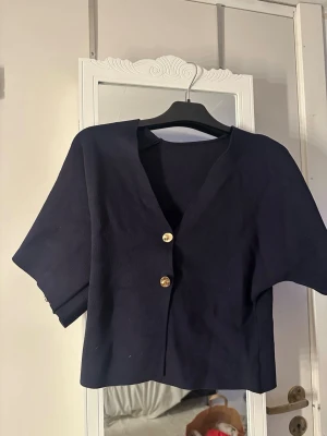 Mörkblå croppad topp med knappar - Trendig mörkblå croppad topp med v-ringning och två guldfärgade knappar framtill. Toppen har korta ärmar och en stilren, minimalistisk design som passar perfekt till jeans eller kjol. tröjan är i storlek S 