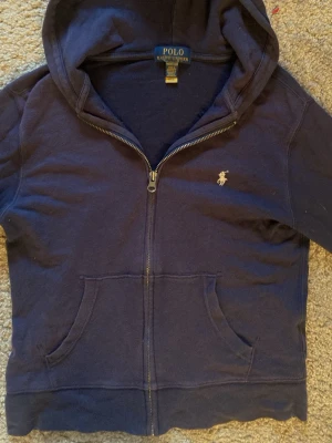 Marinblå zip hoodie från Polo Ralph Lauren - Klassisk marinblå hoodie från Polo Ralph Lauren med dragkedja framtill och broderad logga på bröstet. Hoodien har känguruficka och mjukt material som känns skönt mot huden. Perfekt för en chill och stilren look. Super skön och är en liten tunnare variant, men passar till alla årstider. Den är köpt för 1500, Ge gärna prisförslag!!💕