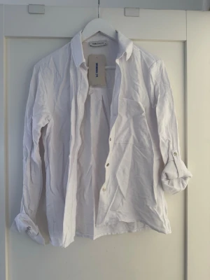 Vit skjorta från LC Waikiki - Klassisk vit skjorta från LC Waikiki i bomull med lång ärm och knappar framtill. Skjortan har en bröstficka och enkel krage, perfekt för en clean och stilren look. Ärmarna kan rullas upp och fästas med knapp.