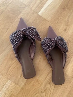 Skor  - Säljer ett par bruna mules från H&M med spetsig tå och stora rosetter täckta av pärlor. Mulesen har öppen häl och en elegant design som sticker ut. Perfekta för dig som gillar detaljer och vill ha något unikt på fötterna.