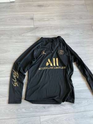 Svart PSG träningströja Jordan - Snygg svart långärmad träningströja från PSG och Jordan med guldfärgade detaljer och loggor. Halvlång dragkedja framtill och texten 'Paris' på ena ärmen. Tillverkad i ett lätt och funktionellt material som passar perfekt till träning.