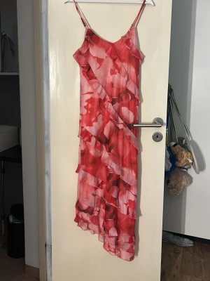 Rosa och röd blommig långklänning - Säljer en somrig långklänning med tunna axelband och blommigt mönster i rosa och rött. Klänningen har volangdetaljer och är tillverkad i ett lätt, luftigt material som passar perfekt för varma dagar. Aldrig använd