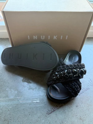 Svarta sandaler från Inuikii med pärlor - Säljer ett par svarta sandaler från Inuikii med breda, stickade band dekorerade med svarta pärlor och stenar. Sandalerna har en chunky sula och öppen tå, vilket ger en trendig och avslappnad look. Perfekta för dig som vill ha något unikt och bekvämt till sommaren. 