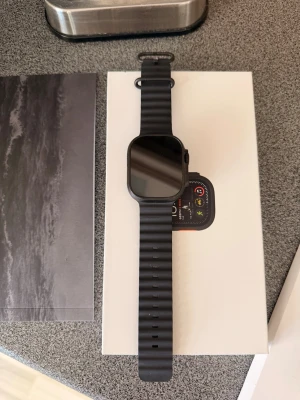 Apple watch ultra 2 - Snygg svart apple watch med fyrkantig display och svart silikonarmband. Klockan har en modern design med rundade hörn och flera sensorer på baksidan för hälsomätning. Perfekt för dig som vill ha koll på träning och notiser direkt på handleden. Den är i väldigt gott skick. Inga repor, smuts som syns på bild