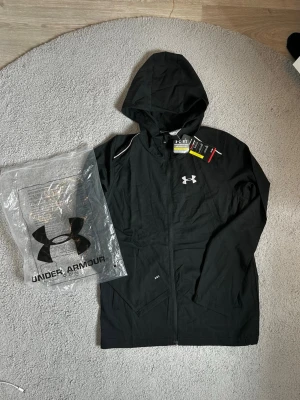 Svart hoodie från Under Armour - Snygg svart hoodie från Under Armour med dragkedja och huva. Logga på bröstet och stor text på ryggen. Tillverkad i lätt och funktionellt material, perfekt för träning eller chill. Fickor på sidorna och diskreta detaljer vid axlarna.
