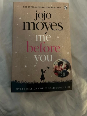 Me Before You av Jojo Moyes - En internationell bästsäljare av Jojo Moyes. Pocketbok med beige omslag och svart text, inslag av rosa och vitt. Omslaget har en siluett av en kvinna och stjärnor, samt en filmrelaterad sticker. Romanen handlar om Lou Clark och Will Traynor och deras känslosamma resa.
