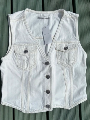 Vit jeansväst från Abercrombie & Fitch - Snygg vit jeansväst från Abercrombie & Fitch med fyra knappar framtill, två bröstfickor med lock och knapp samt justerbart spänne i ryggen. Västen har en klassisk v-ringad form och är gjord i kraftigt bomullstyg. Perfekt för att lyfta din outfit.
