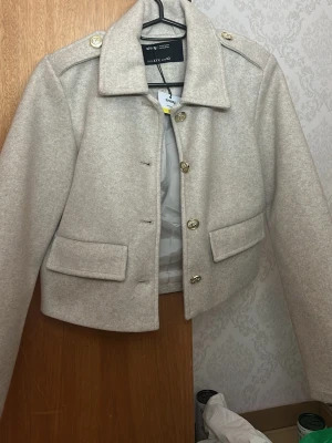 Ljusbeige ullkappa  - Kort, ljusbeige ullkappa med guldknappar och två stora fickor framtill. Klassisk krage och rak passform ger en stilren look. Perfekt för dig som vill ha en trendig och elegant jacka till hösten.