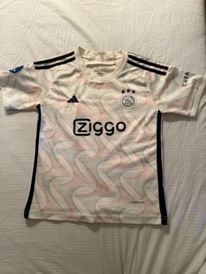 Ajax matchtröja  - Ajax matchtröja i vitt med grafiskt mönster i pastellfärger, svarta detaljer och tre stjärnor ovanför klubbmärket. Ziggo-logga på bröstet, Tadić och nummer 10 på ryggen. Tillverkad i lätt och ventilerande Aeroready-material. Korta ärmar och klassiska Adidas-ränder på axlarna. Finns en liten fläck som säkert går bort i tvätten. Passar 160-165cm