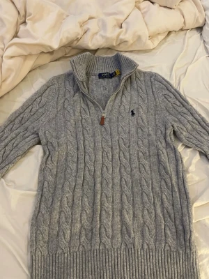 Grå kabelstickad tröja Polo Ralph Lauren - Kabelstickad grå tröja från Polo Ralph Lauren med halv dragkedja och hög krage. Tröjan har ribbade muddar och nederkant samt den klassiska loggan broderad på bröstet. Aldrig använd då jag fick den men den sitter riktigt bra o de finns inga defekter