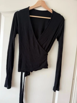 Svart omlottopp med långa ärmar - Stilren svart omlottopp med långa ärmar och knytband i midjan. Djup v-ringning och mjukt, stretchigt material som sitter snyggt på kroppen. Perfekt att matcha med jeans eller kjol för en trendig look.