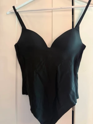 Svart body med smala axelband - En helt ny Snygg svart body med vadderade kupor och smala justerbara axelband. Den är aldrig använd. Tillverkad i ett stretchigt och mjukt material som sitter tight mot kroppen. Perfekt att styla under skjortor eller tröjor för en clean look.