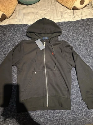 Svart hoodie från Polo Ralph Lauren - Svart hoodie med dragkedja från Polo Ralph Lauren. Klassisk design med huva, dragsnören och kängurufickor. Liten röd broderad logga på bröstet. Tillverkad i mjukt bomullsmaterial för en skön känsla.