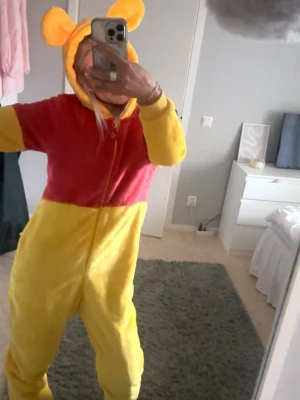 Nalle Puh  - Gul och röd onesie inspirerad av Nalle Puh med huva och söta öron. Dragkedja framtill och mjukt, fluffigt material som är perfekt för maskerad eller cosplay. Passar dig som vill sticka ut och vara extra mysig på temafesten!