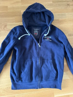 Marinblå hoodie från Morris - Snygg marinblå hoodie från Morris med dragkedja, fickor framtill och vit dragsko i huvan. Broderad logga och text på bröstet. Tillverkad i mjukt bomullsmaterial som känns skönt mot huden. Perfekt för en avslappnad och stilren look.