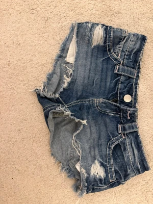 Blå slitna jeansshorts med fransar - Säljer ett par blå jeansshorts med slitna detaljer och fransig kant. Shortsen har synliga sömmar, fem fickor och dekorativa nitar på bakfickorna. Perfekta för sommaren och ger en avslappnad vibe.