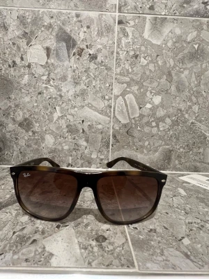  solglasögon från Ray-Ban - Snygga svarta solglasögon från Ray-Ban med klassisk fyrkantig form och mörka tonade glas. Bågarna är i plast och har Ray-Ban-loggan på sidan. Perfekta för soliga dagar och ger en cool vibe till din outfit.