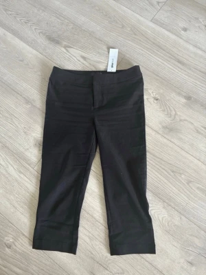 Capri byxor Gina Tricot - Stilrena svarta kostymbyxor med croppad benlängd och klassisk passform. Byxorna har en enkel midja utan synliga fickor och stängs med dragkedja och hake. Perfekta för en clean och modern look. Endast provade. 