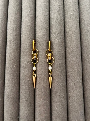 Guldpläterade örhängen med pärldetalj - Snygga guldpläterade örhängen med små vita pärlor och spetsiga hängen. Örhängena har cirkelformade detaljer och gnistrande stenar på öglan. Perfekta för dig som gillar en edgy men elegant stil.