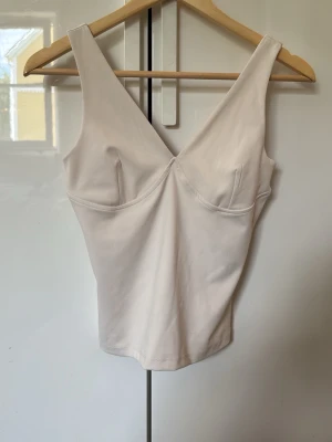 Beige croppad linnetopp med v-ringning - Snygg vit linnetopp med breda axelband och markerad v-ringning både fram och bak. Toppen är croppad och har en figurnära passform som framhäver siluetten. Perfekt att styla med jeans eller kjol för en clean look.